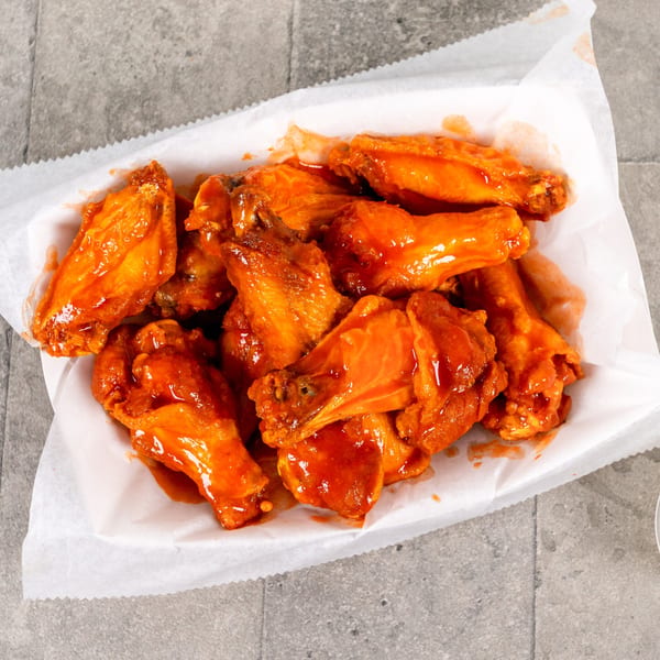 A white paper-lined basket filled with numerous bright orange, saucy chicken wings on a gray surface.