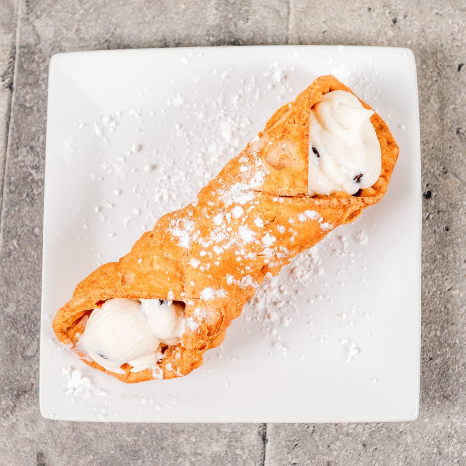 A golden-brown cannoli filled with white cream and chocolate chips, dusted with powdered sugar.