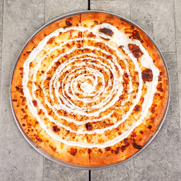 Overhead view of a round pizza with white sauce drizzled in a spiral pattern and chicken.