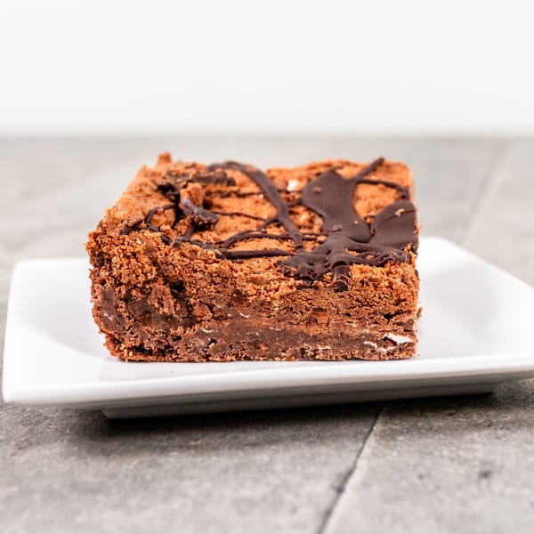 A square chocolate brownie with a chocolate drizzle on a white rectangular plate.