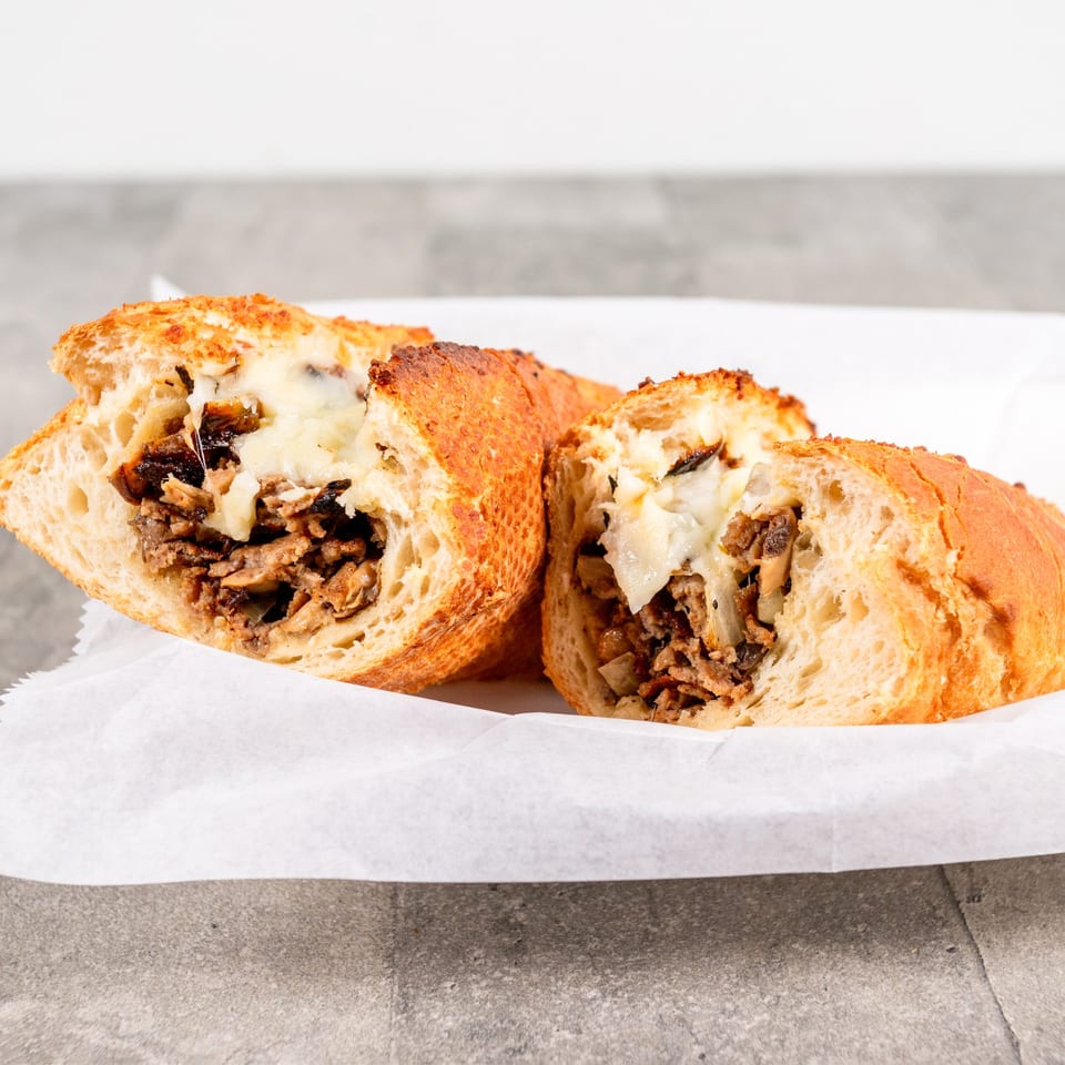 Two halves of a cheesy steak and mushroom sandwich on a white paper-lined tray.